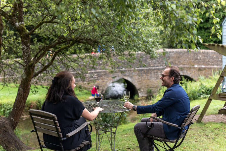 A man and woman drinking wine at a table with the bridge in Ford, Wiltshire in the background